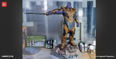 Thanos 3D model in armor with double-bladed weapon, surrounded by paints and supplies.