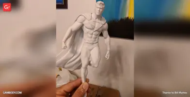 3D-printed model of a muscular superhero in a cape, posed mid-flight, held in someone's hand.