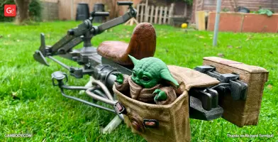 A 3D-printed speeder bike with a small green alien figure in a tan pouch on a grassy lawn.