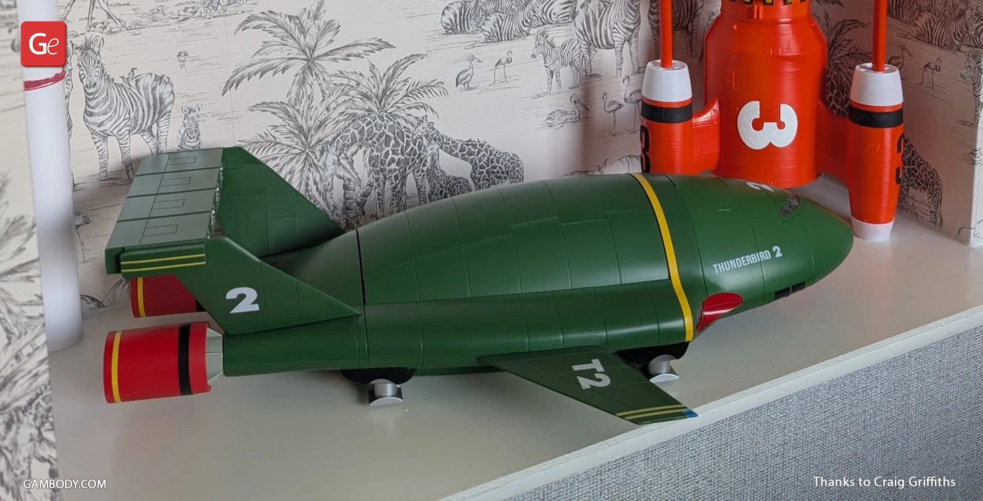Green Thunderbird 2 model with red accents, showcased on a shelf against illustrated wallpaper.