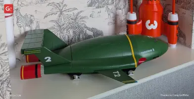 Green Thunderbird 2 model with red accents, showcased on a shelf against illustrated wallpaper.