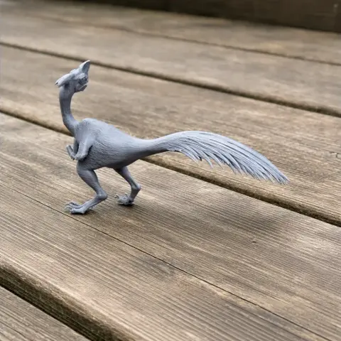 3D printed gray Oviraptor model with a long tail and crest, displayed on a wooden surface.