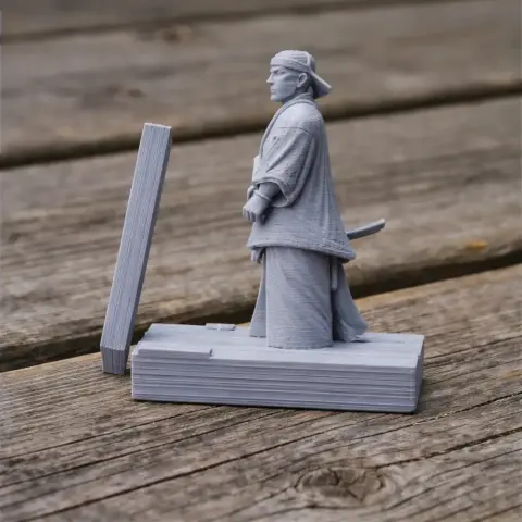 3D-printed samurai figure in traditional attire, standing on a wooden base outdoors.