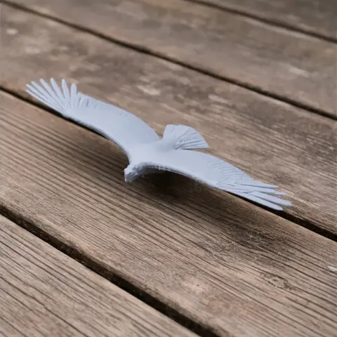 Gray 3D printed model of a bird in flight, wings spread, resting on a wooden surface.