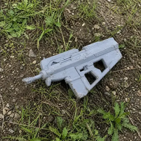A gray 3D-printed sci-fi prop gun lies on grass and soil.