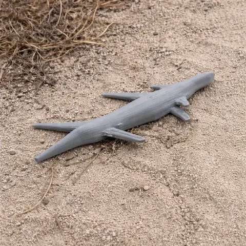 A small, gray 3D printed dinosaur or marine reptile figurine lying on sandy ground.