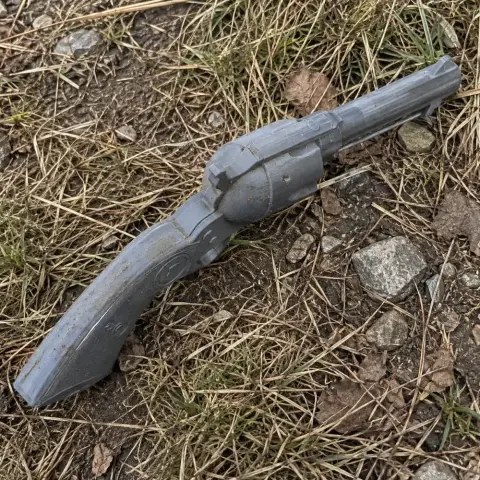 3D printed gray Colt revolver prop lying on grass and dirt.