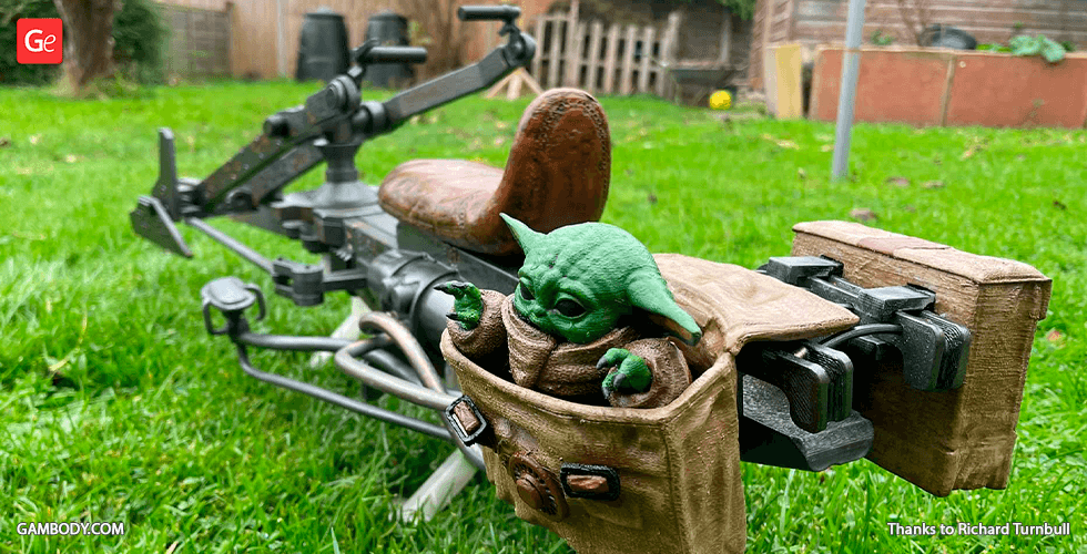 A 3D-printed speeder bike with a small green alien figure in a tan pouch on a grassy lawn.