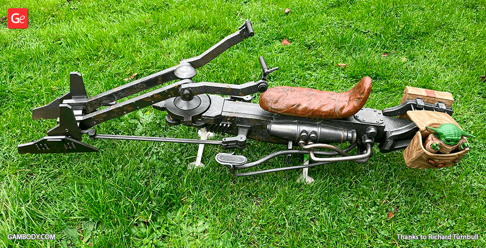 A detailed 3D-printed Mandalorian speeder bike on grass with a small green character in a satchel.